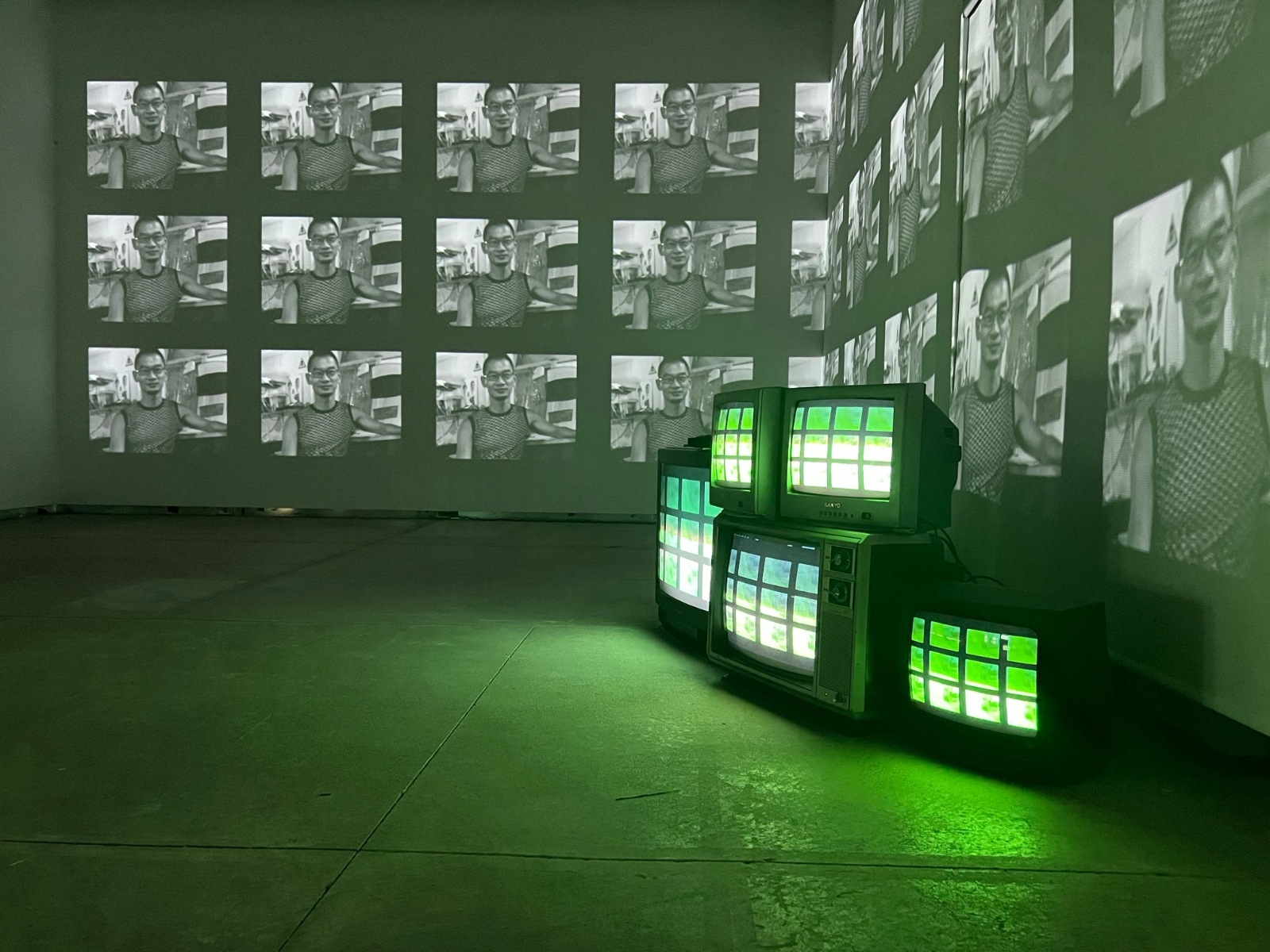 Dimly-lit room with five retro TVs stacked together playing green visuals. The room is empty otherwise, with portrait projections shown on the walls.